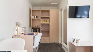 A compact hotel room with a desk, chair, microwave, and a TV mounted on the wall.