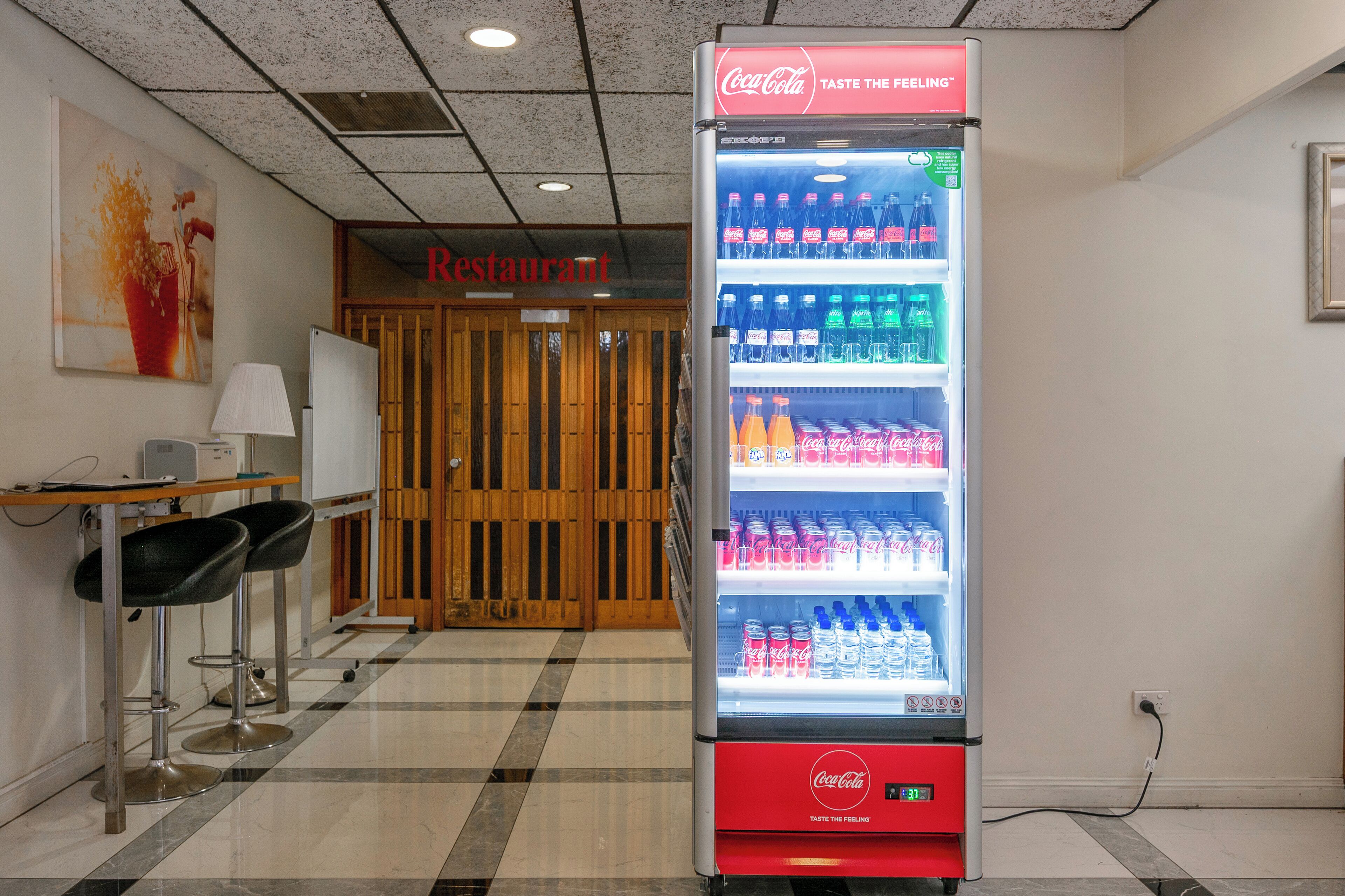 A vending machine stocked with Coca-Cola products in a room with a checkered floor, a whiteboard, and two black bar stools.
