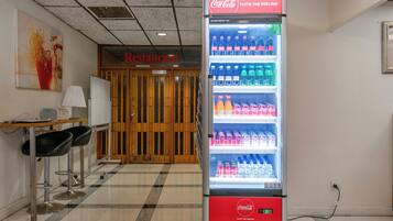 A vending machine stocked with Coca-Cola products in a room with a checkered floor, a whiteboard, and two black bar stools.
