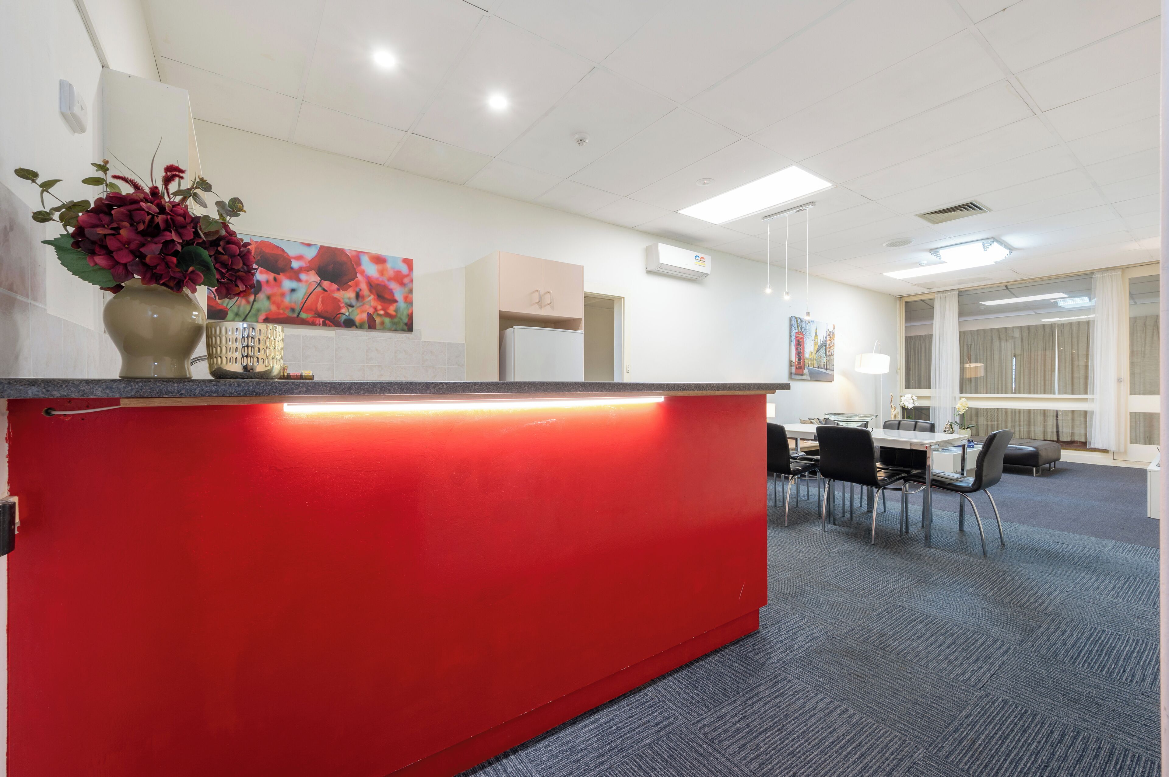 A reception area with a red counter, a vase with flowers, a painting on the wall, and a dining area with tables and chairs.