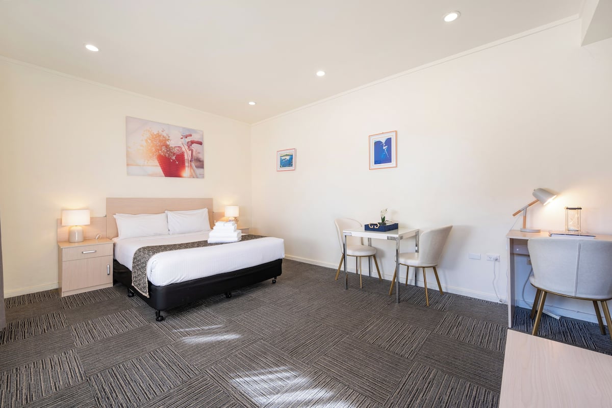 A hotel room with a bed, bedside tables, a desk with a chair, and artwork on the walls.