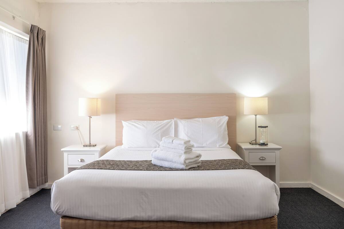A neatly made bed with white linens and a folded towel, two bedside lamps, a nightstand, and a window with curtains.