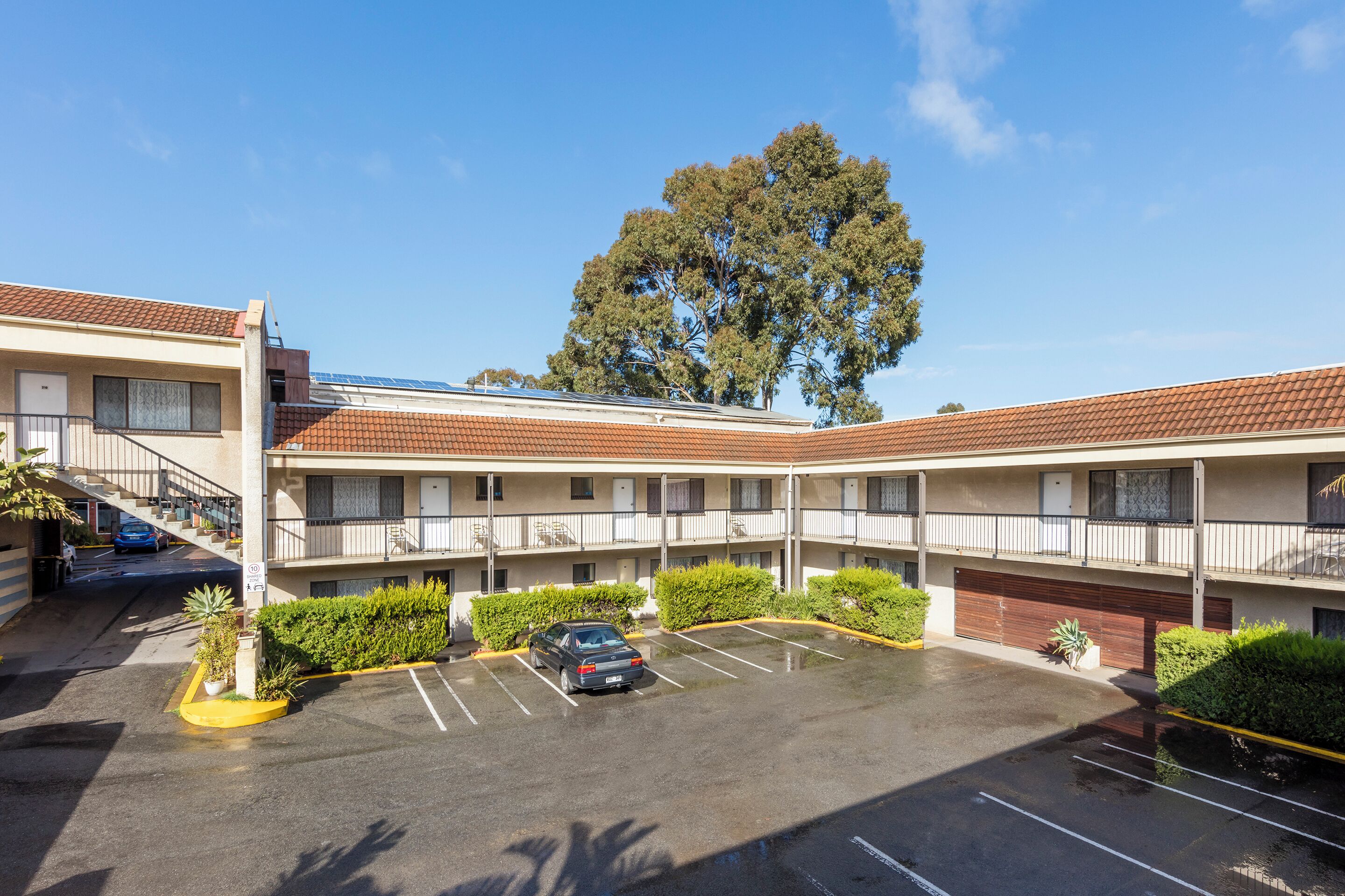 Ein mehrstöckiges Motel mit Parkplatz, überdachtem Gang und einem Baum im Hintergrund.