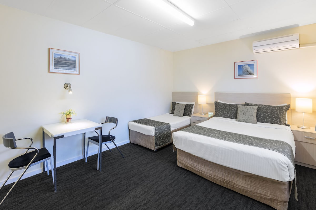 A hotel room with two single beds, a desk with a chair, a small table with a vase, and two wall-mounted lamps.
