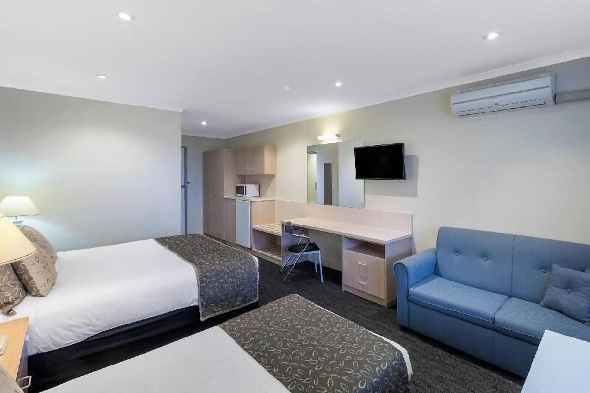 A hotel room with two beds, a desk, a chair, a blue sofa, a flat-screen TV, and an air conditioning unit.