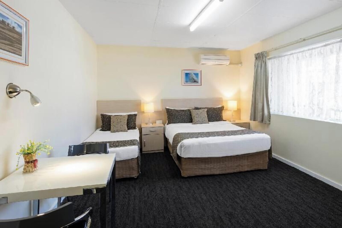 A hotel room with a bed, a desk, a chair, a nightstand, and a window with curtains.