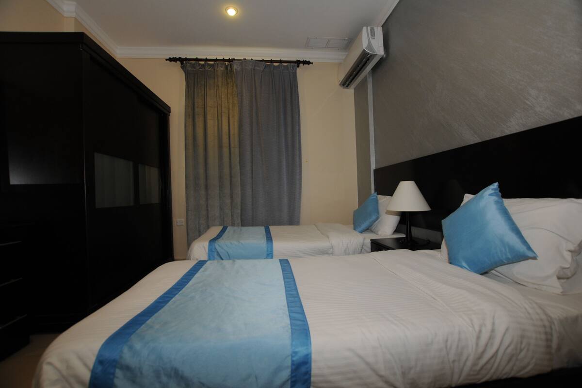 A hotel room with two beds, a dark wardrobe, a window with grey curtains, a white lamp, and an air conditioning unit on the wall.