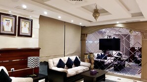 A modern living room with a sofa, armchair, coffee table, and a TV. There are framed pictures on the wall and a decorative lamp. - Al Muhanna Plaza Hotel (Salmiya)