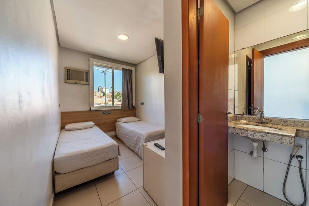 Een kleine hotelkamer met twee eenpersoonsbedden, een bureau, een badkamer met wastafel en spiegel, en een raam met gordijnen.