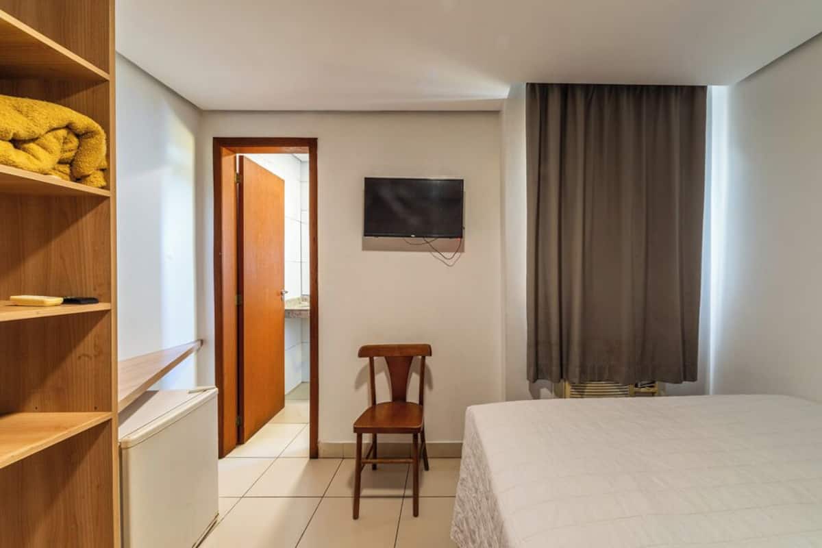 Een hotelkamer met een bed, een aan de muur bevestigde televisie, een houten stoel en een kledingkast met planken.