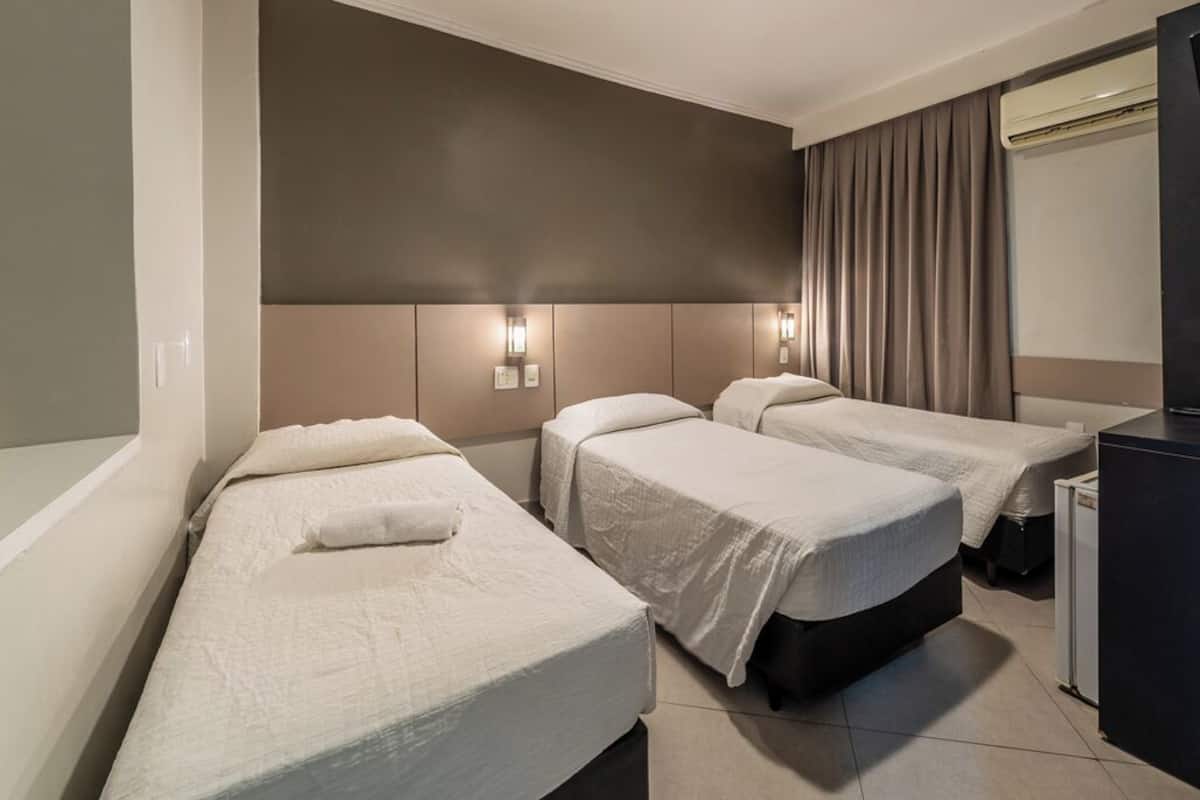 Habitación de hotel con dos camas individuales, un amplio ventanal con cortinas, un escritorio empotrado y un aire acondicionado de pared.