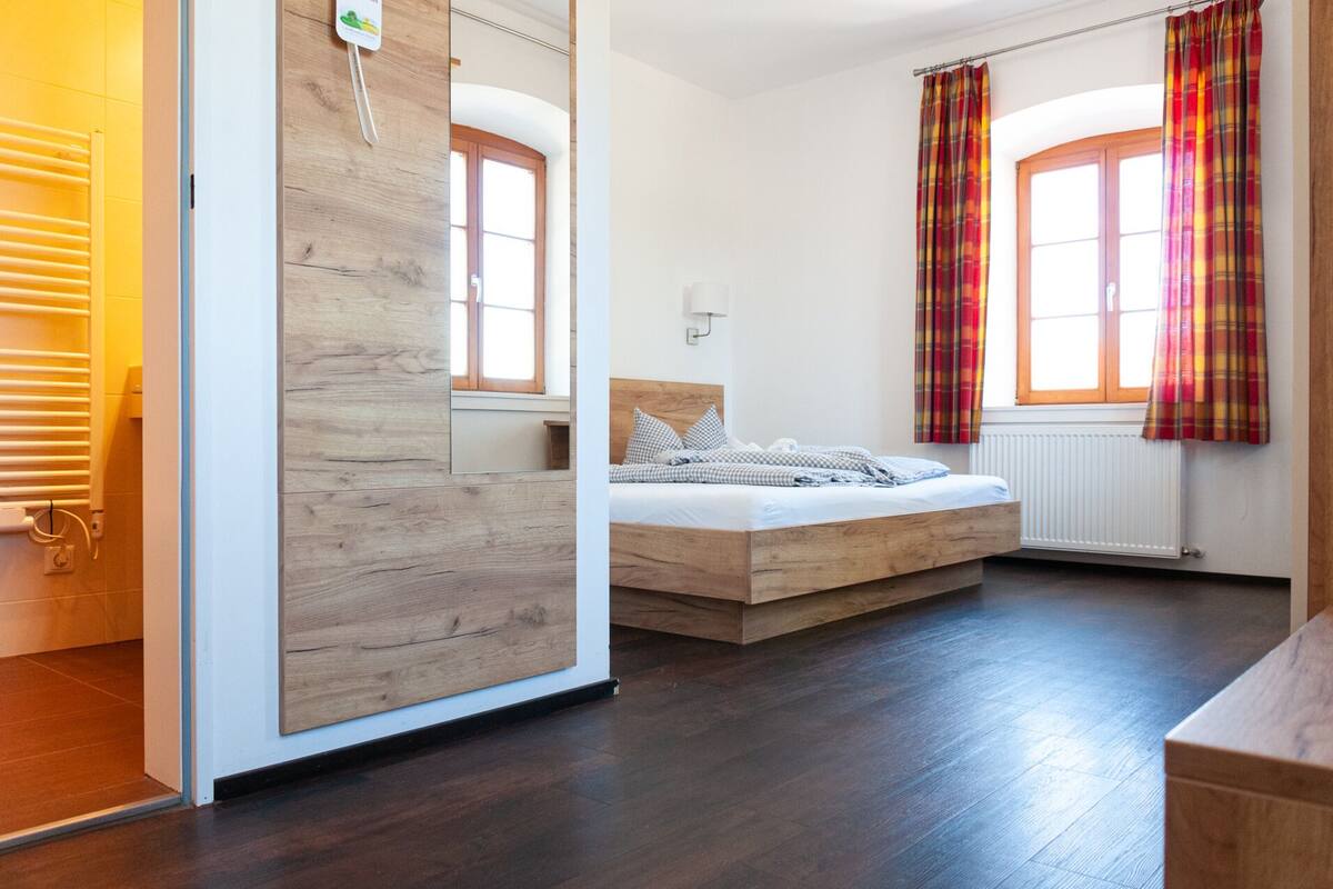 Une chambre avec un plancher en bois, un lit avec une tête de lit, une fenêtre avec des rideaux et un radiateur.