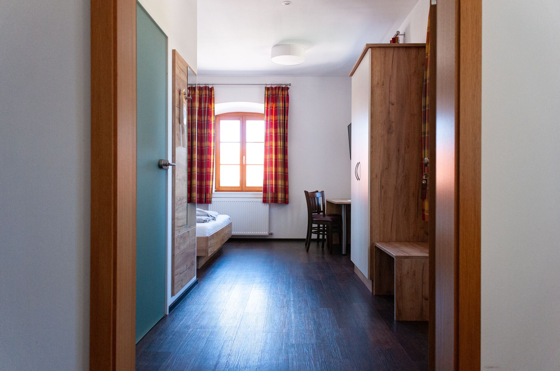 A narrow hallway with a wooden door on the left, a window with plaid curtains, a bed, a wooden desk, and a wardrobe.