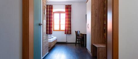 A narrow hallway with a wooden door on the left, a window with plaid curtains, a bed, a wooden desk, and a wardrobe.