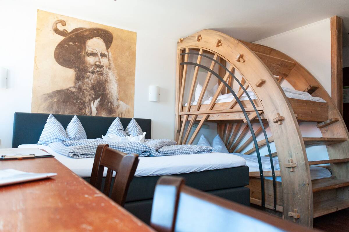 Une pièce comprenant un lit, un lit superposé en bois, une table à manger et un grand portrait accroché au mur.
