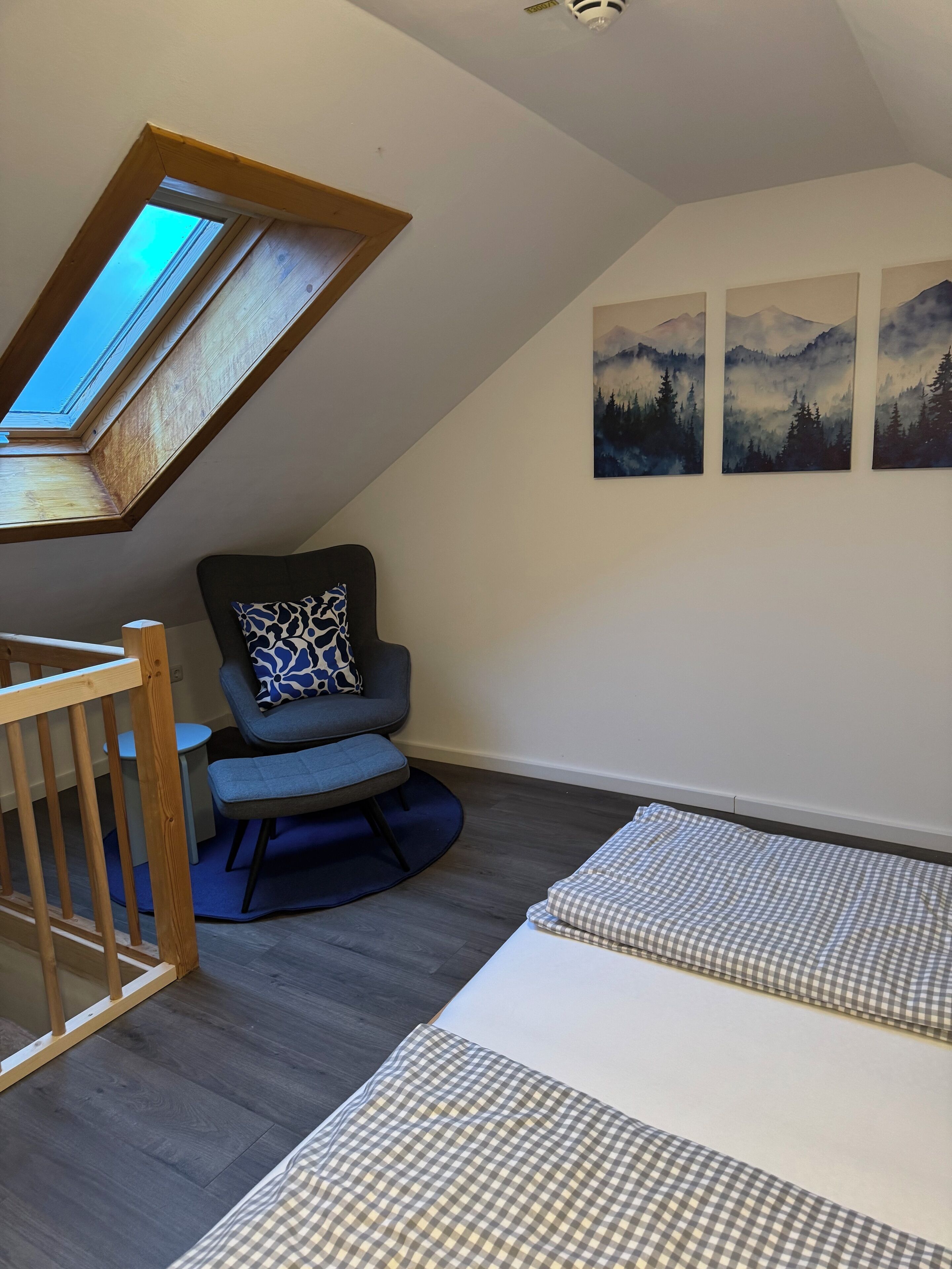 A room with a sloped ceiling, a skylight, a rocking chair, a bed with checkered bedding, and three wall-mounted paintings of mountains.