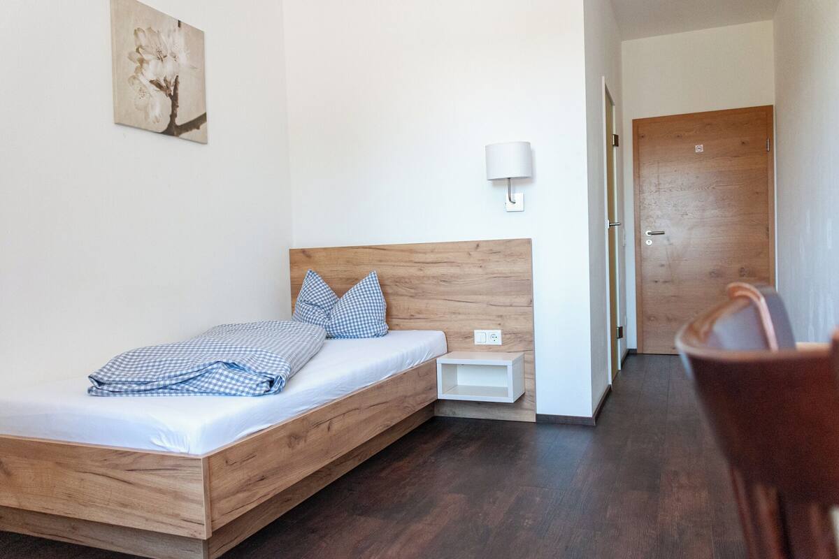 Une chambre avec un lit en bois, une literie à carreaux, une table de chevet, une commode, une lampe fixée au mur et une porte visible au fond.