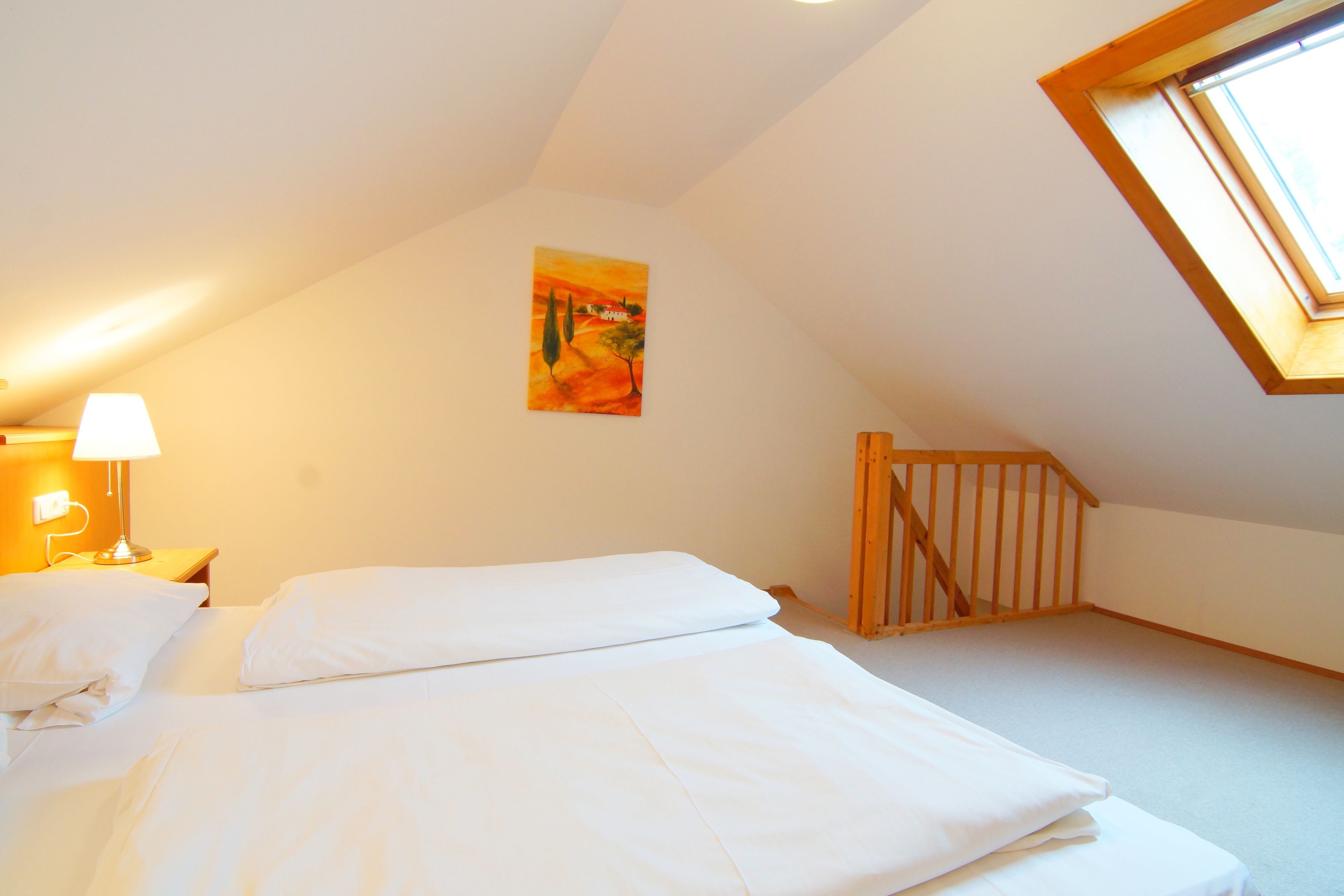 Un dormitorio con techo inclinado, un cuadro en la pared, una escalera de madera y una cama con ropa blanca.