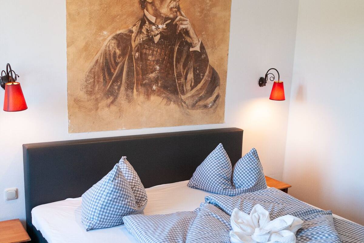 Une chambre d’hôtel avec un grand portrait accroché au mur, un lit recouvert d’une couette à carreaux, deux tables de chevet avec des lampes rouges et une petite table en bois près du lit.