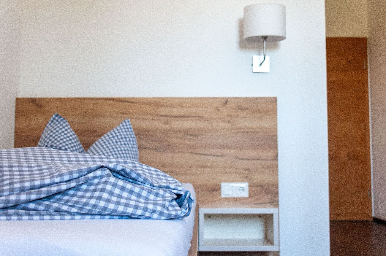 A bedroom with a wooden headboard, a bed with checkered bedding, a bedside table, and a wall-mounted lamp.