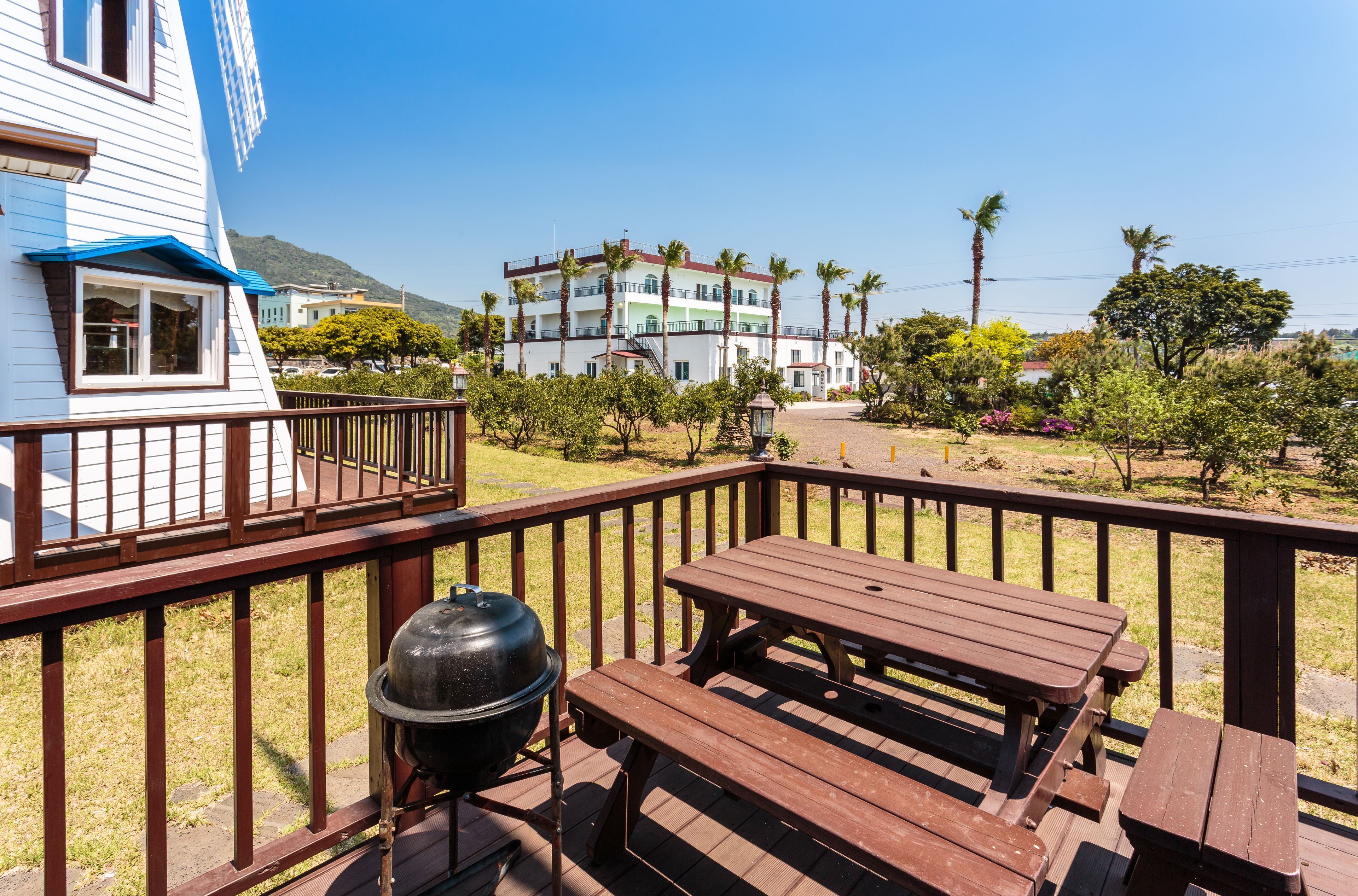 Um deck de madeira com mesas de piquenique e churrasqueira, com vista para uma área arborizada com palmeiras e um edifício ao fundo.