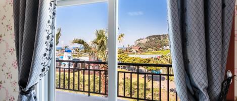 A window with a view of a balcony, a railing, and a distant landscape with buildings and greenery.
