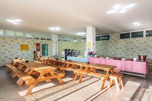 Une grande salle à manger avec des tables et des bancs de pique-nique en bois, un coin cuisine avec une nappe à carreaux rouges et blancs, et un sol carrelé.