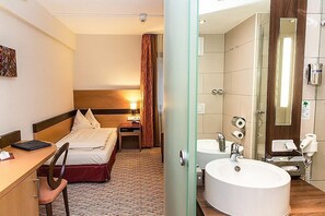 A hotel room with a bed, desk, chair, and a bathroom with a sink and mirror. - TaunusTagungsHotel (Friedrichsdorf)