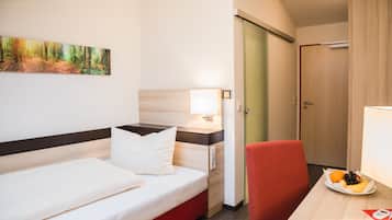 A hotel room with a bed, a desk with a plate of fruit, a red chair, and a painting on the wall.