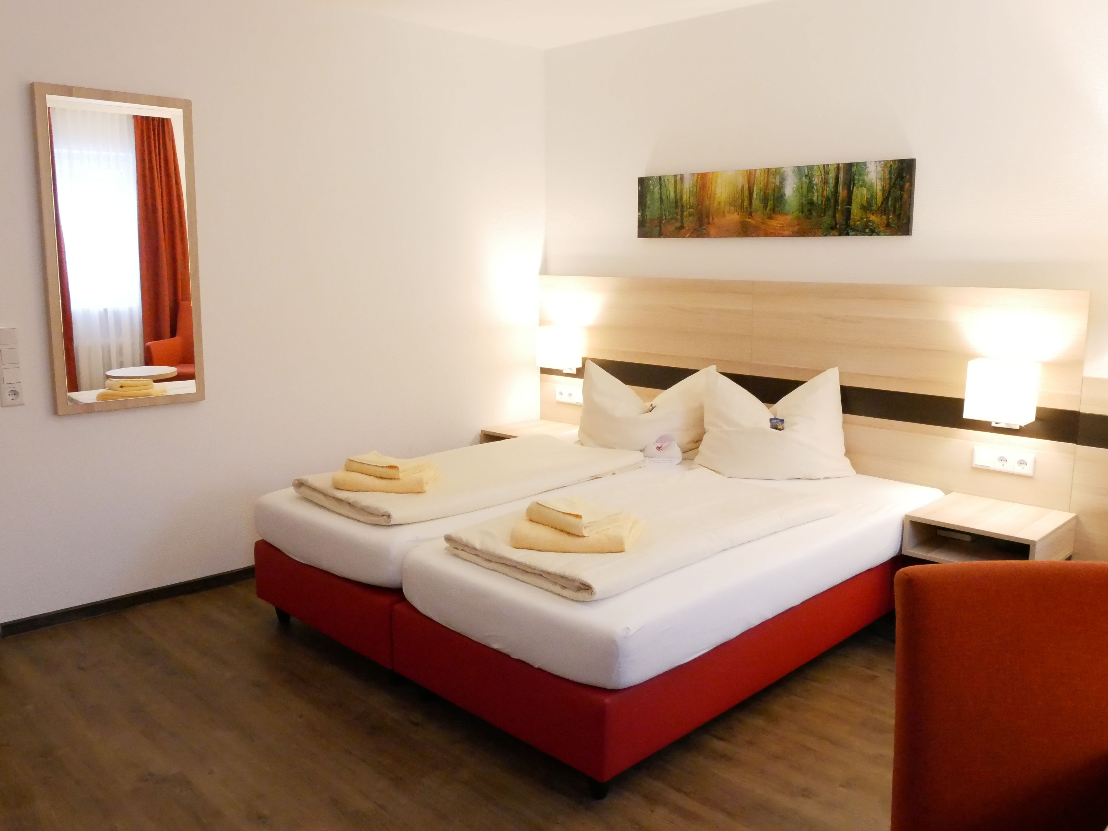 Une chambre d’hôtel avec un lit double, deux tables de chevet, une tête de lit ornée d’un tableau et un fauteuil rouge.