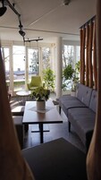 Una sala de estar moderna con un sofá gris, una mesa redonda de centro con un jarrón blanco, una silla verde y grandes ventanales que ofrecen vistas al exterior.