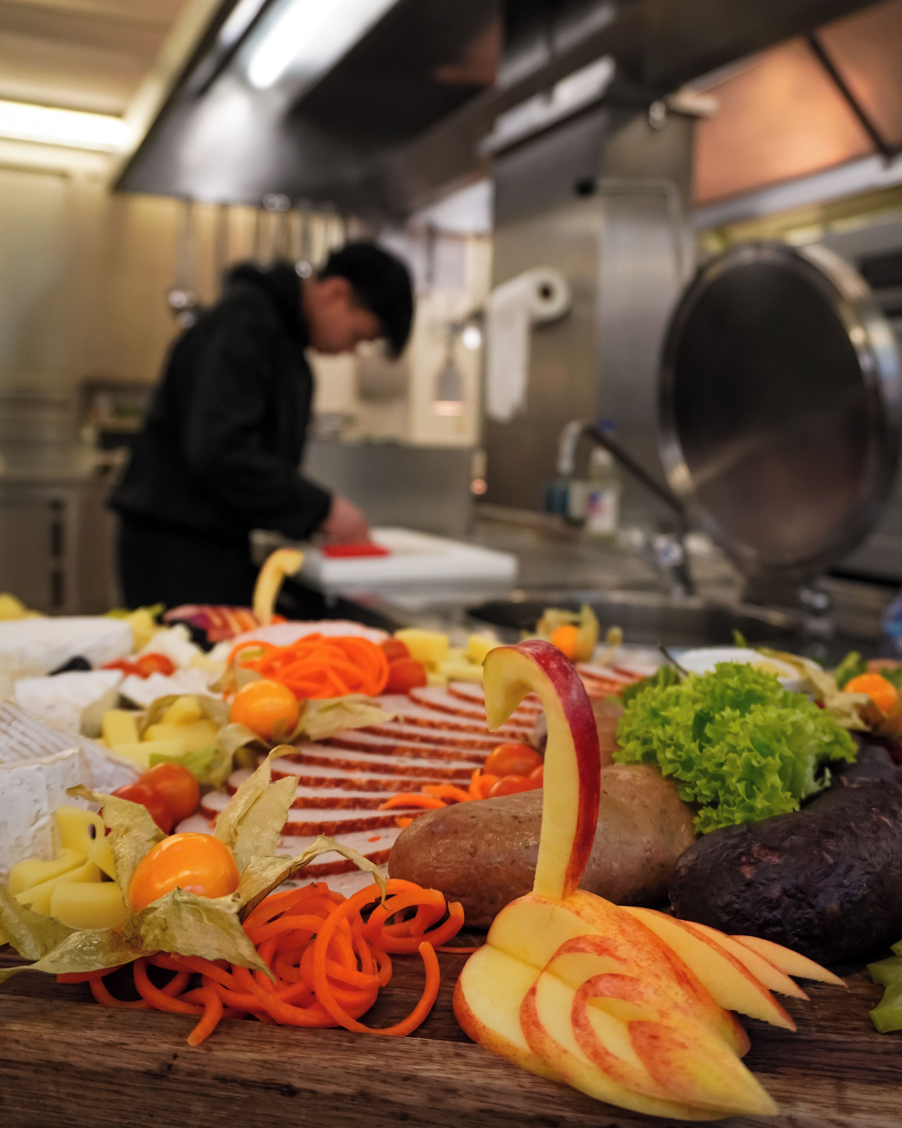 Una cocina con un chef preparando comida, una variedad de frutas y verduras frescas en la encimera y electrodomésticos de acero inoxidable al fondo.
