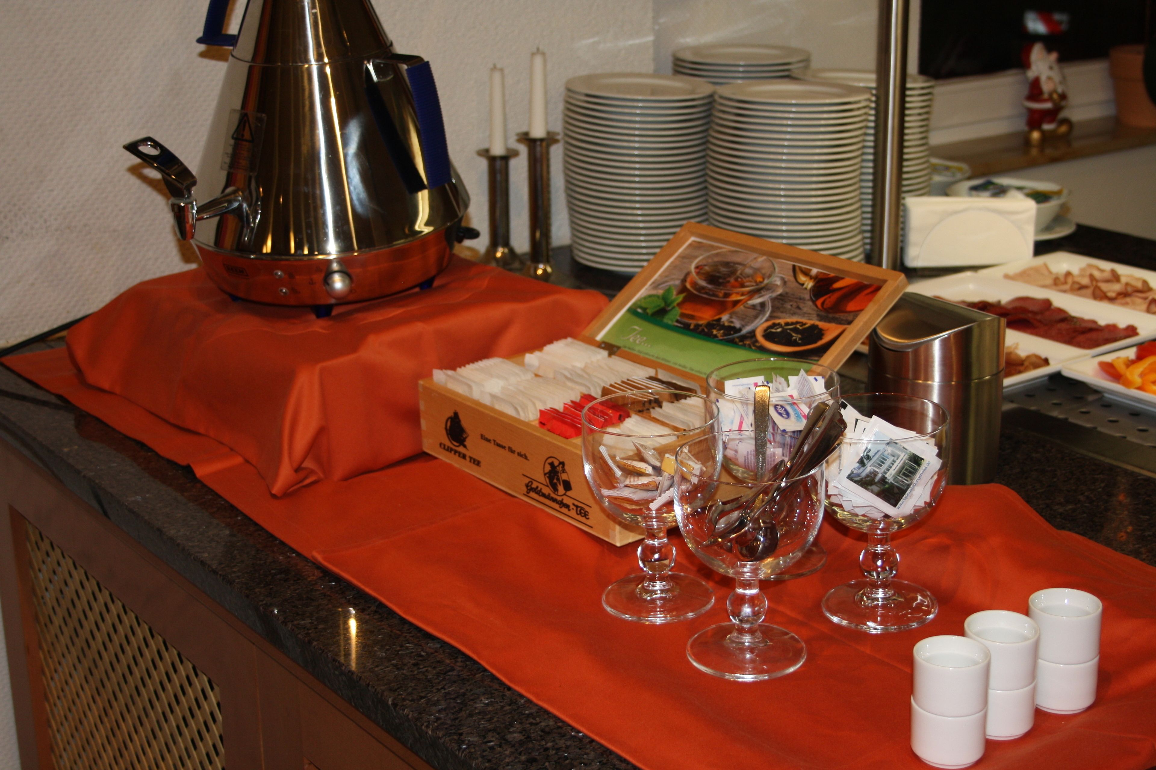 Montaje de bufé con diferentes opciones de comida, una cafetera y varias copas de vino sobre un mantel rojo.