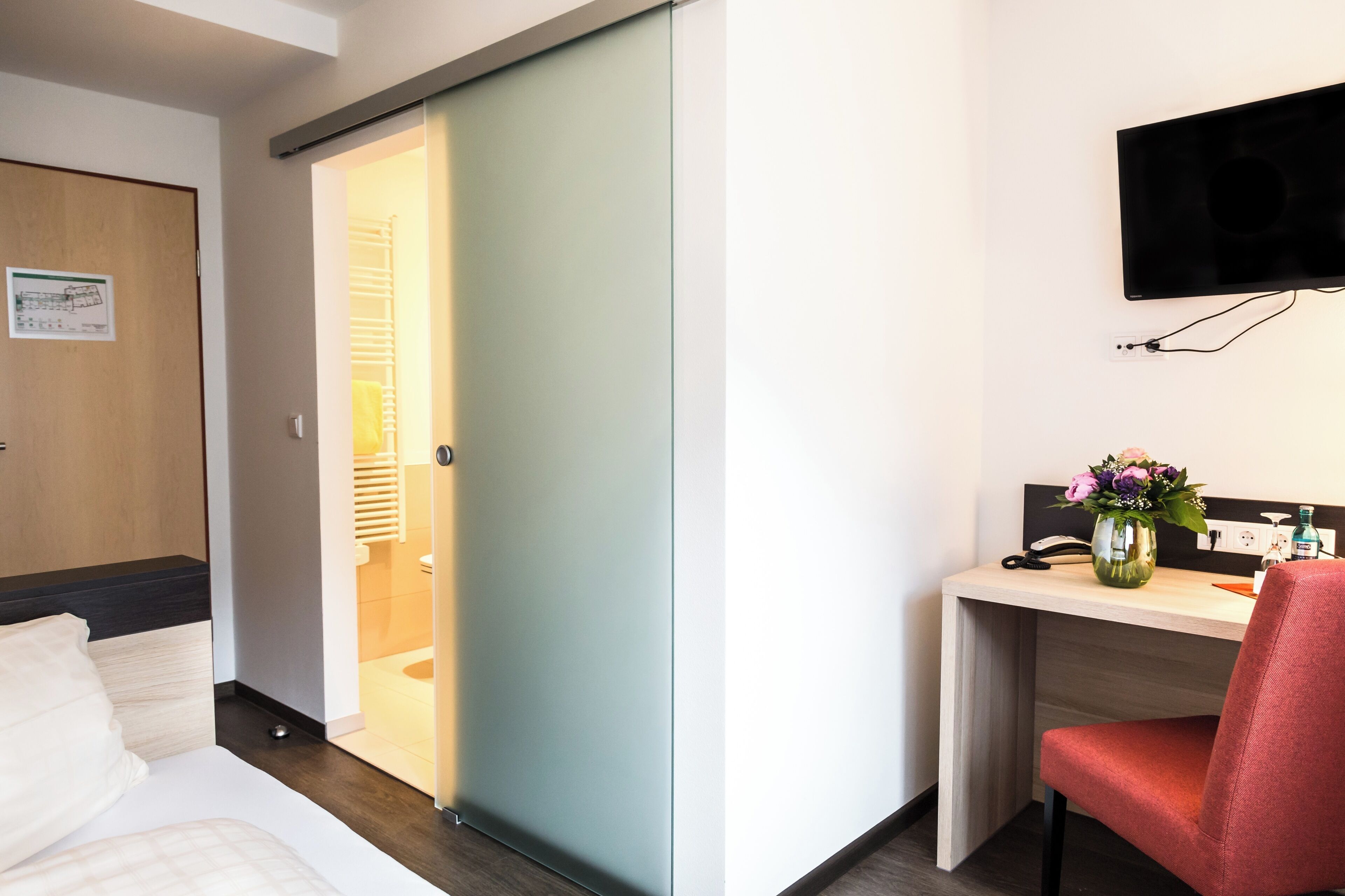A hotel room with a bed, a desk with a chair, a TV mounted on the wall, and a sliding door leading to a bathroom.