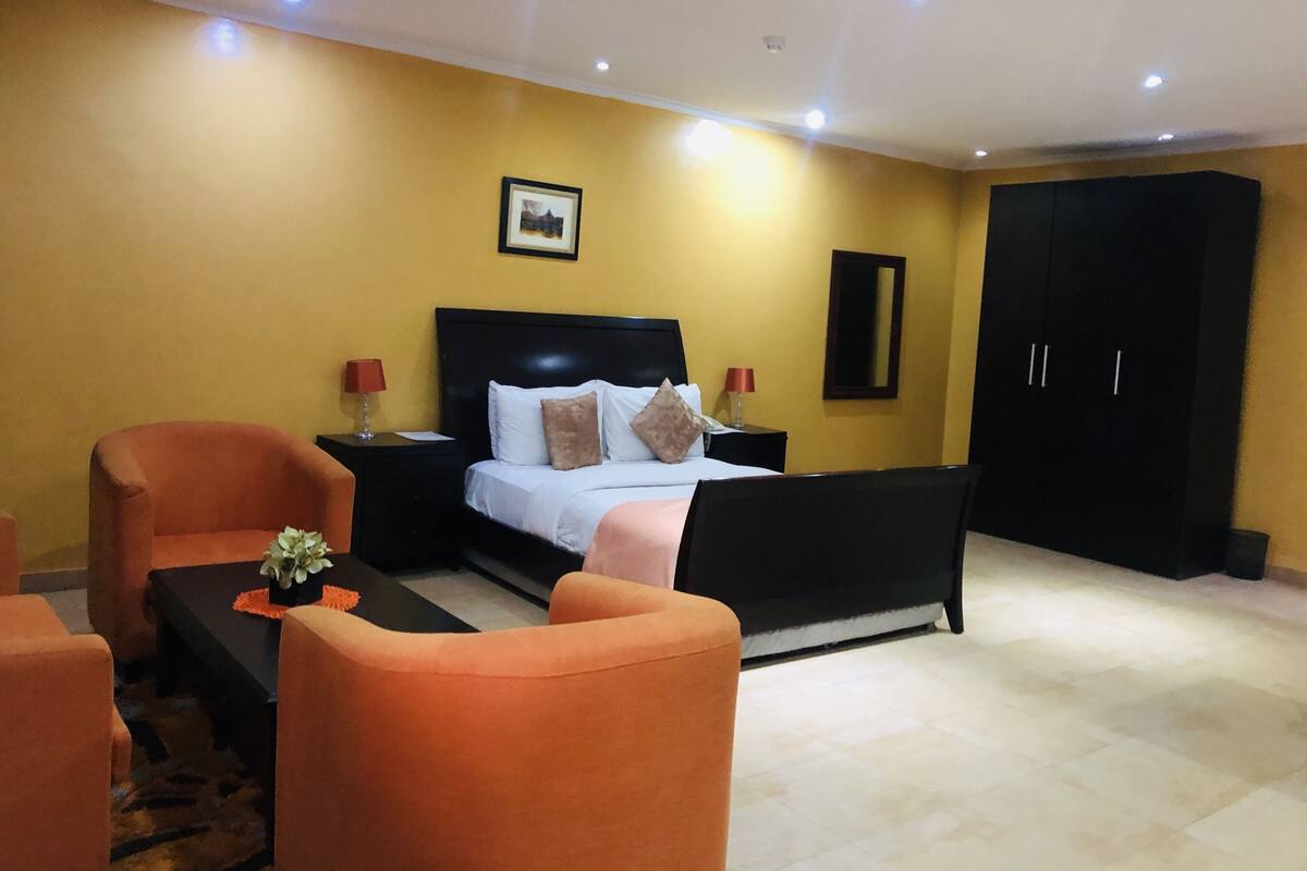 A bedroom with a bed, two nightstands, a black wardrobe, and two orange chairs.
