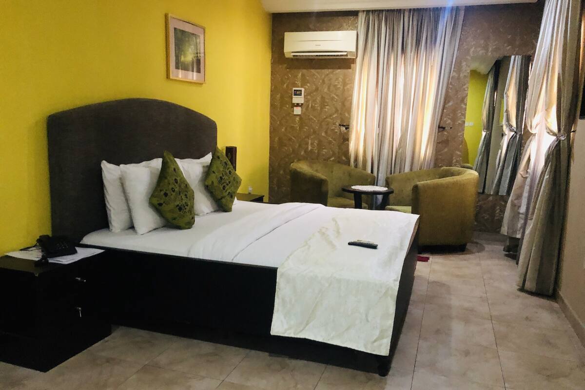 A hotel room with a bed, a nightstand, a chair, an air conditioning unit, and curtains.