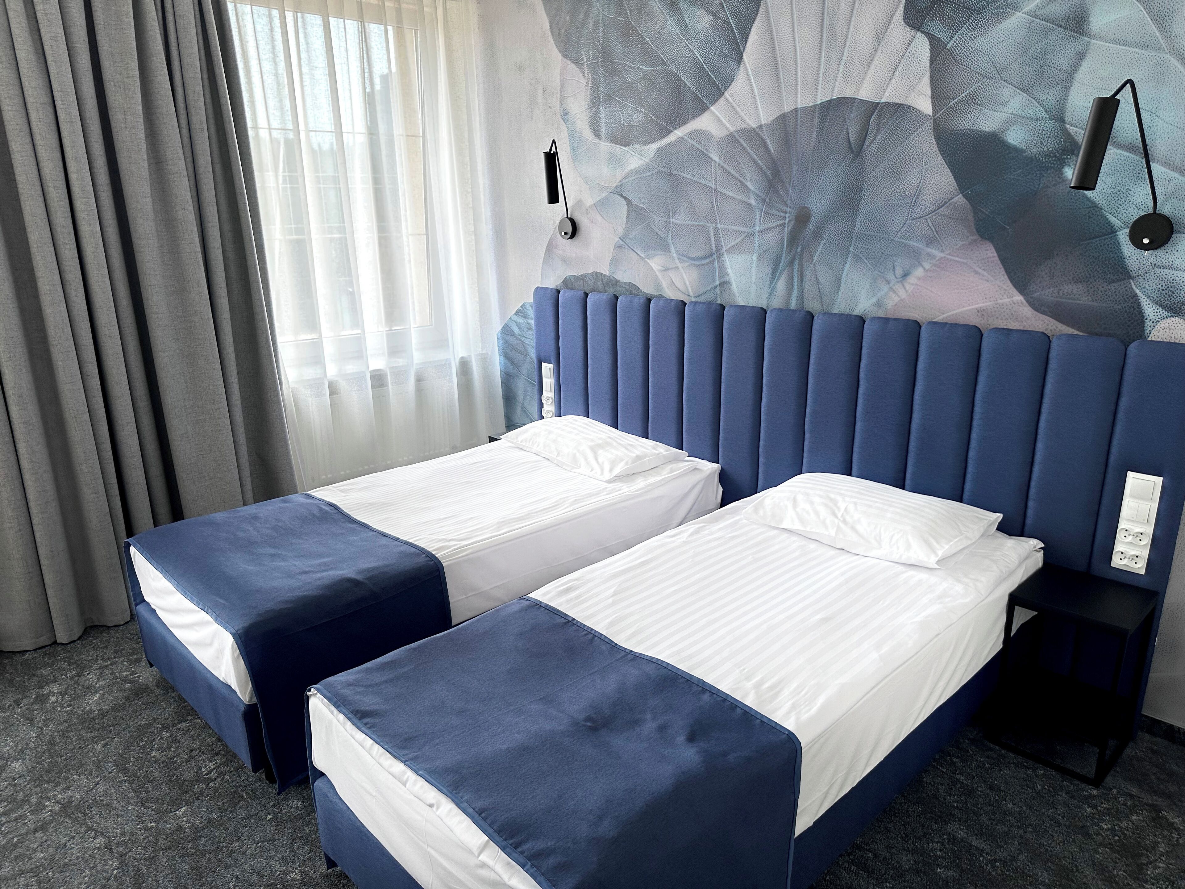 A hotel room with two single beds, a headboard, a bedside table, and a wall-mounted lamp.