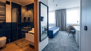 A modern hotel room with a glass-enclosed shower, a separate sink, a bed with a blue headboard, a desk with a chair, and a large mirror.