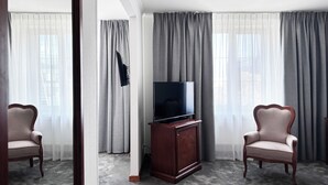 Habitación de hotel con televisión de pantalla plana sobre un mueble de madera, dos sillones tapizados y un ventanal con cortinas grises.