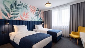 Habitación de hotel con dos camas, un escritorio, una silla, un ventanal grande con cortinas y una pared con diseño de hojas.