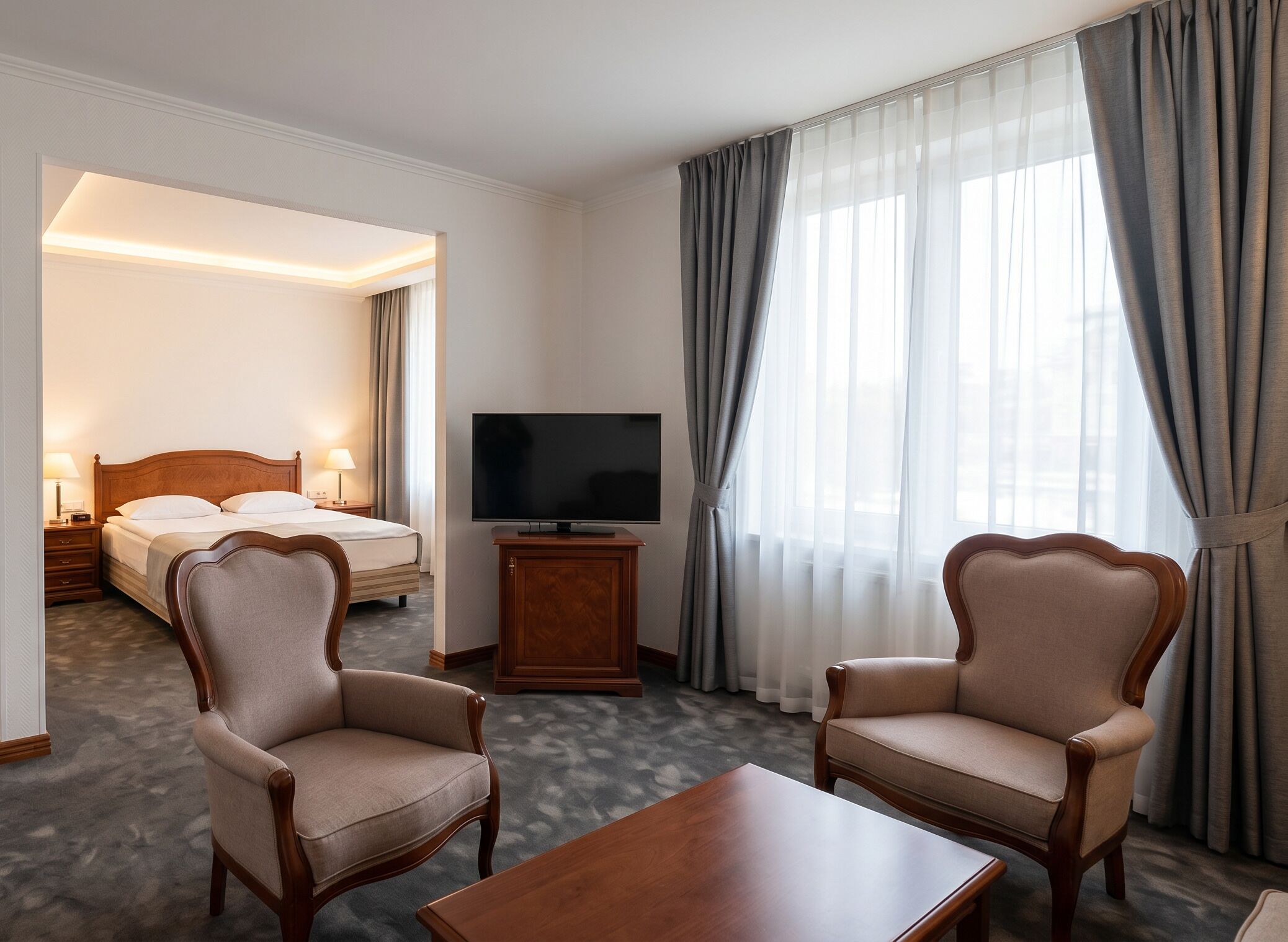 A hotel room with a bed, a TV, two armchairs, a coffee table, and a view of the outside through large windows.