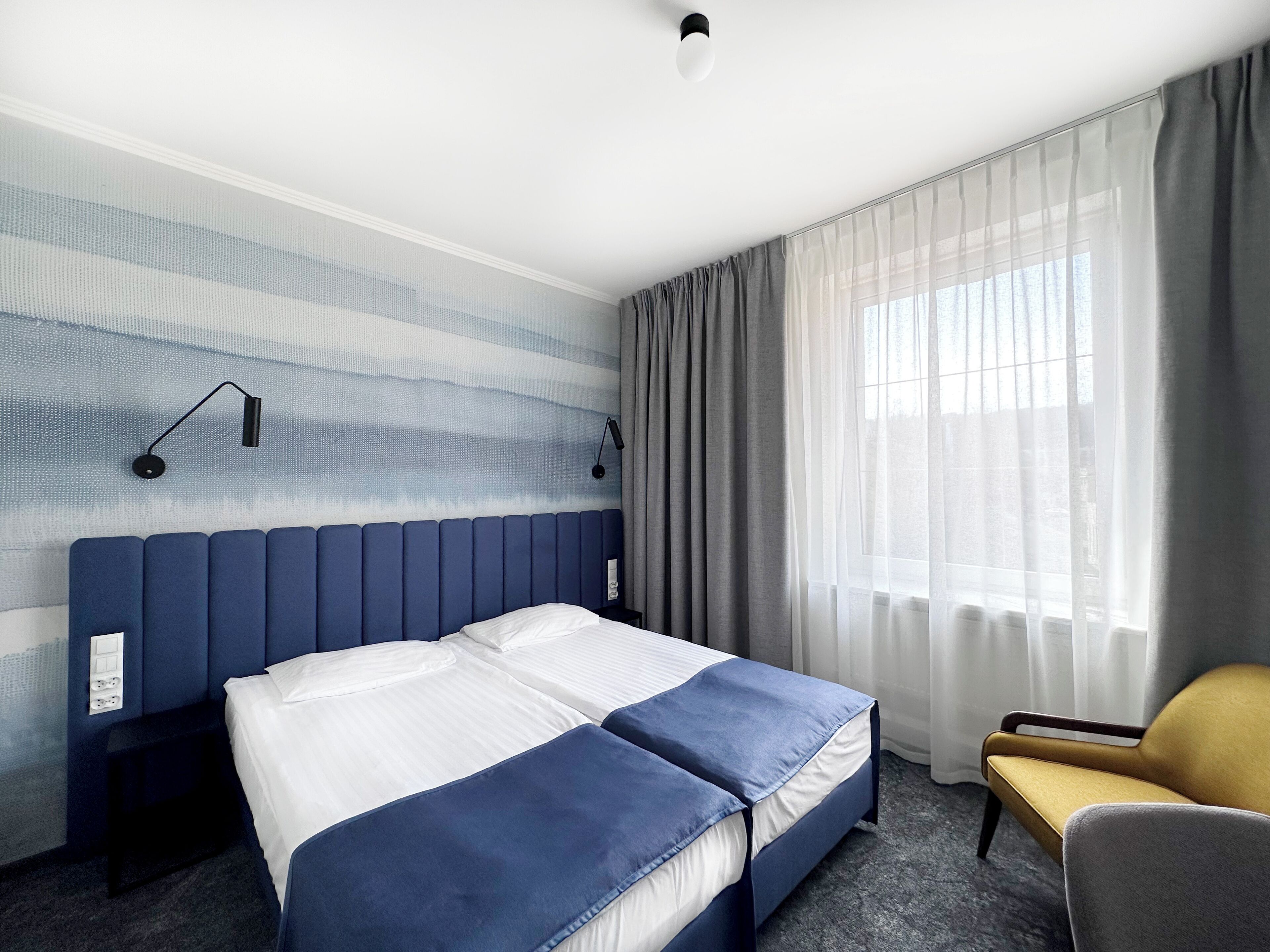A hotel room with a bed, a chair, a window with curtains, and a wall with a textured design.