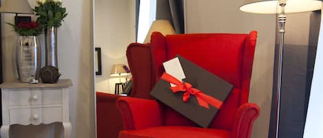 A red armchair with a gift box and magazines, a white side table with a vase and lamps, and a large mirror on the wall.