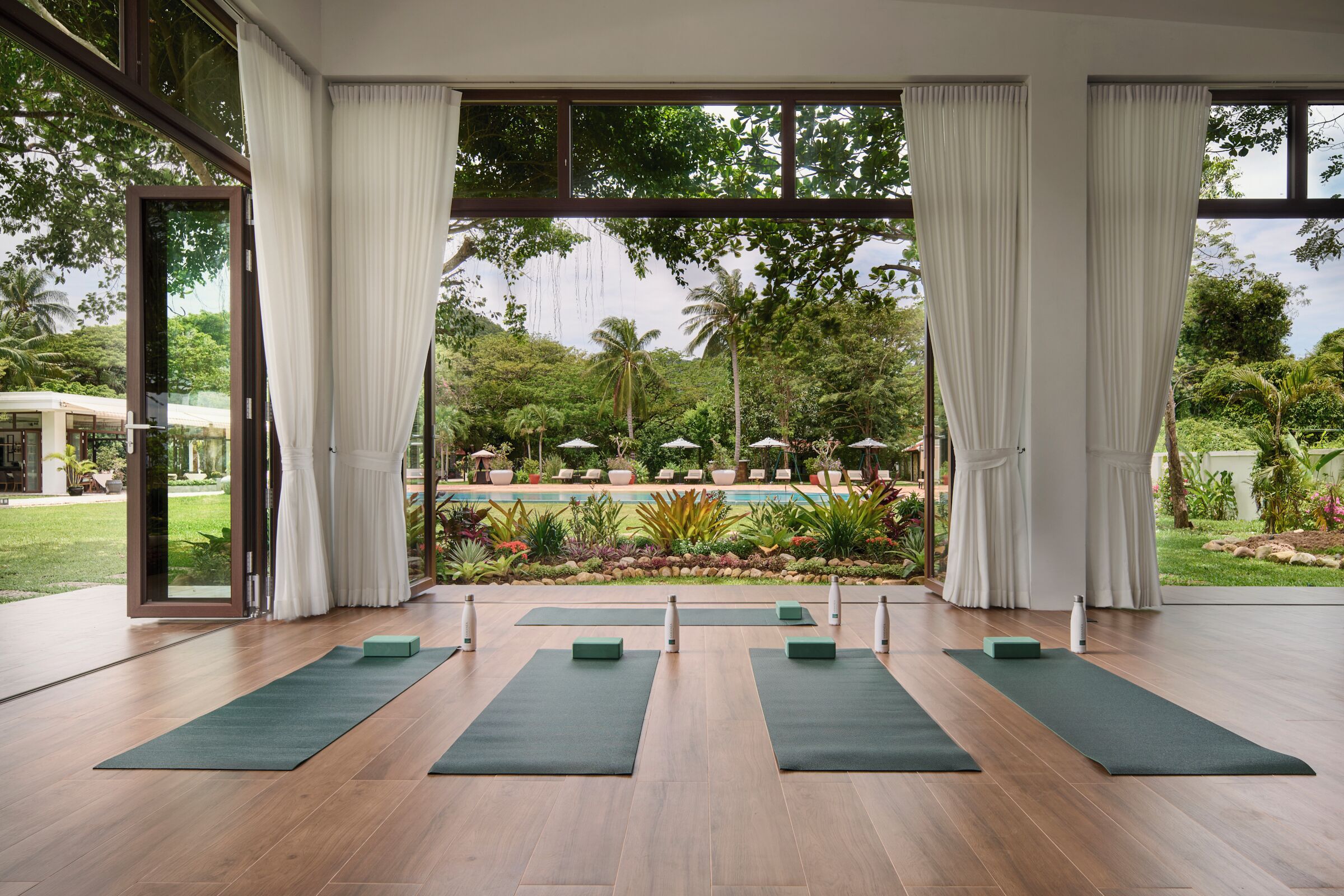 Una sala de yoga con ventanas amplias, suelo de madera y esterillas verdes.