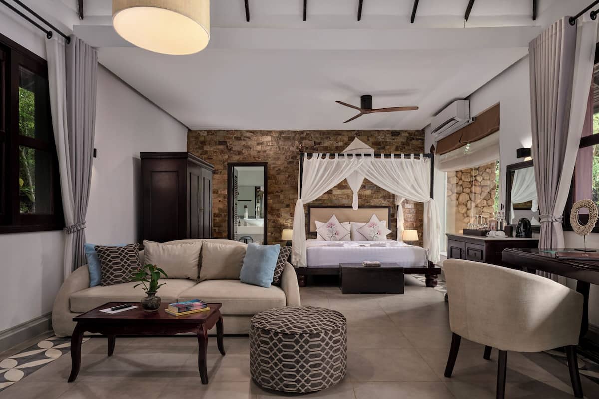 A bedroom with a bed, a sofa, a coffee table, and a chair. There is a mirror on the wall and a ceiling fan.