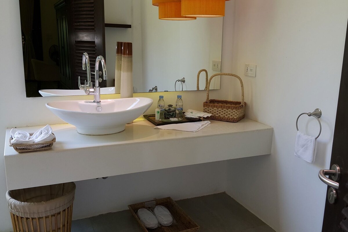 A bathroom with a large white sink, a mirror, a wicker basket, and a small trash bin.