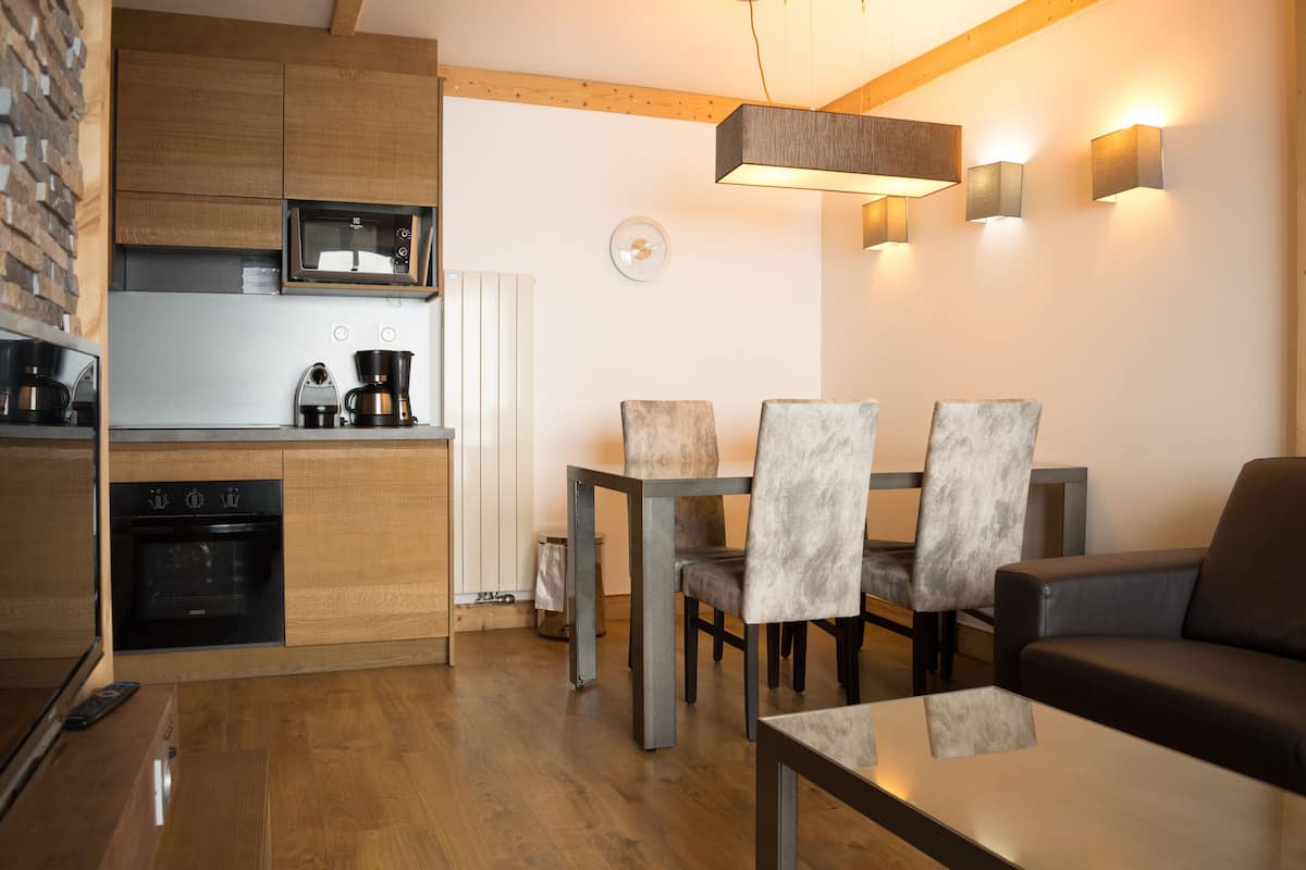 A modern kitchen with wooden cabinets, a built-in oven, and a microwave. There is a dining area with a table and chairs, and a sofa in the living room.