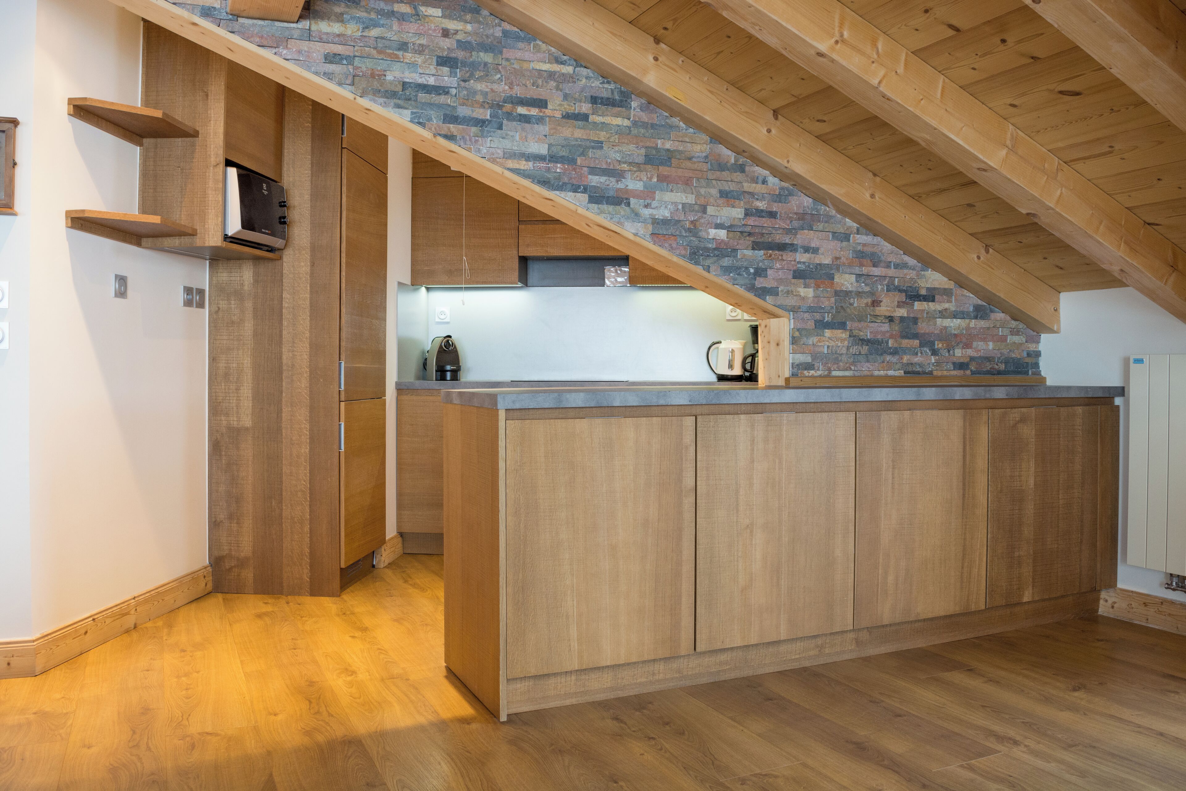 A modern kitchen with wooden cabinets, a brick wall, and a sloped ceiling.