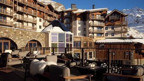 A ski resort with multiple wooden buildings, outdoor seating areas with chairs and tables, and a view of snow-covered mountains. - Résidence Koh-I Nor by Les Etincelles (Les Belleville)