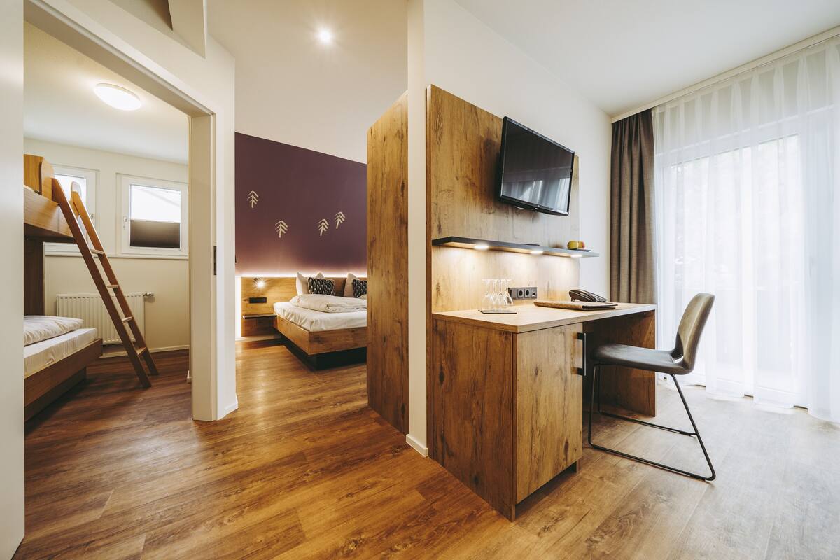 A modern room with a wooden floor, a bed, a desk with a chair, a TV mounted on the wall, and a window with curtains.